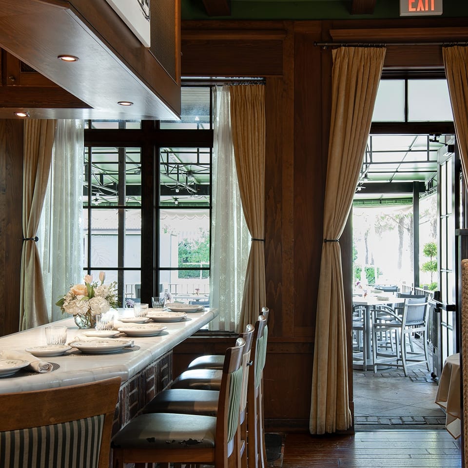 Warmly lit restaurant dining room with wood paneling, set tables, upholstered chairs, tall curtains, and an outdoor patio entrance.