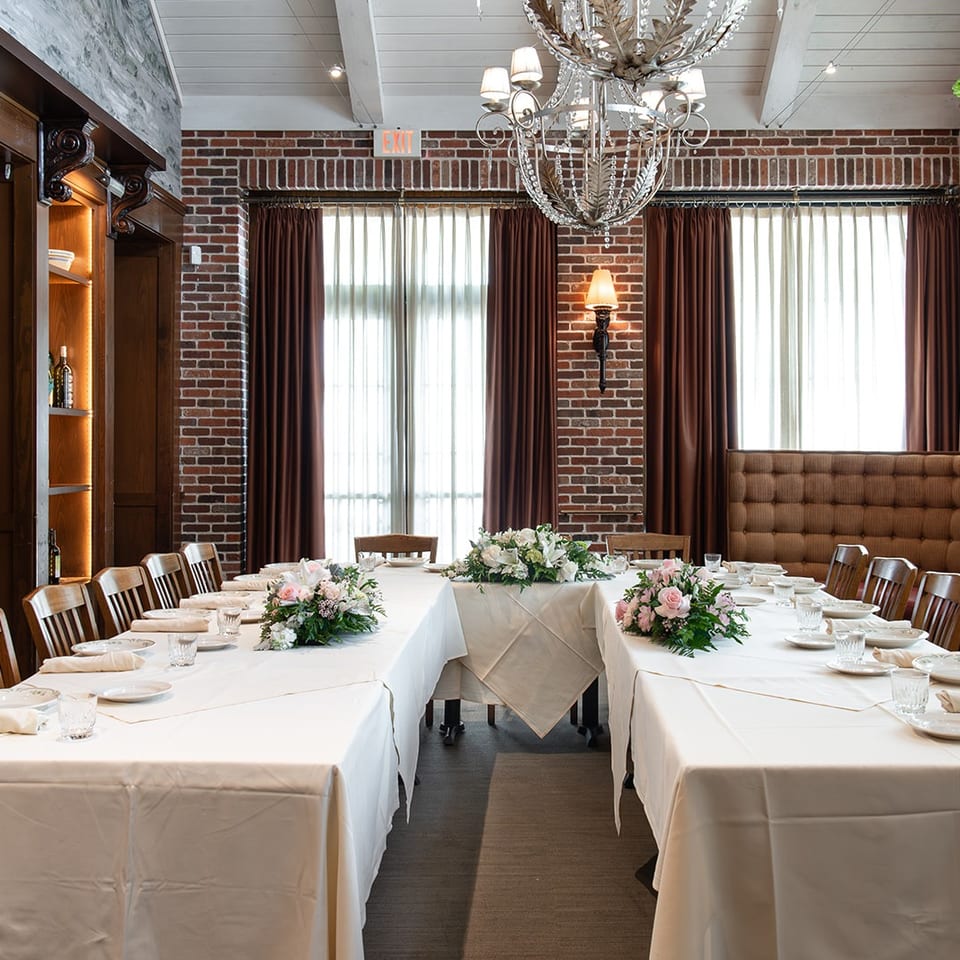 Elegant restaurant dining room with white-clothed tables, floral centerpieces, tufted booth seating, brick walls, and a chandelier.